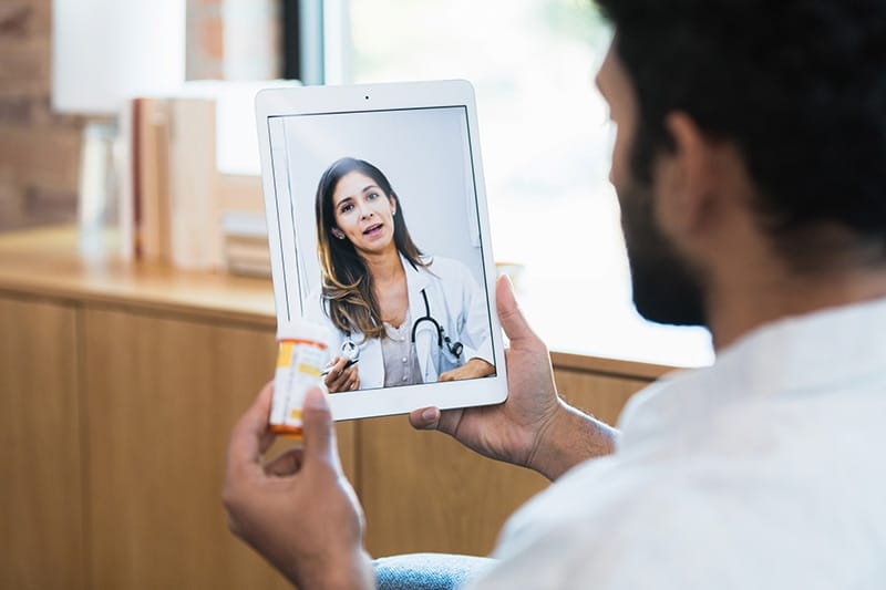 The unrecognizable man listens to the female doctor's advice about the medication he is holding in his hand.  He uses his digital tablet to access a telemedicine app.